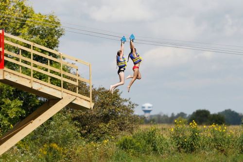 Two people ziplining from a wooden platform over a grassy field under a cloudy sky.