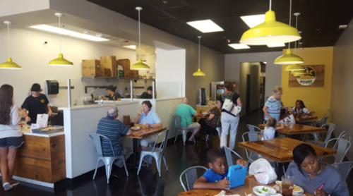 A busy restaurant interior with customers dining, staff serving, and bright yellow pendant lights overhead.