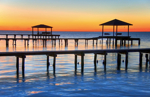 Sunset over calm waters with two pavilions on piers, reflecting vibrant orange and blue hues in the sky.