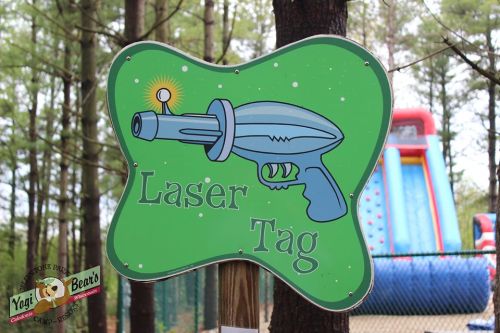 Sign for "Laser Tag" featuring a cartoon laser gun, set in a wooded area with a colorful inflatable in the background.