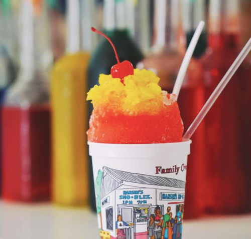 A colorful snow cone topped with a cherry in a cup, with vibrant syrups in the background.