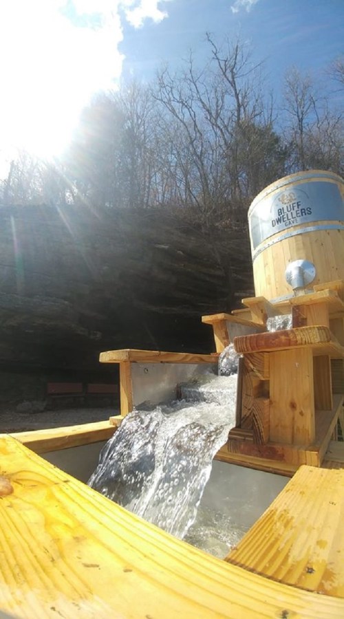 A wooden water feature with flowing water, set against a rocky backdrop and bright sunlight.