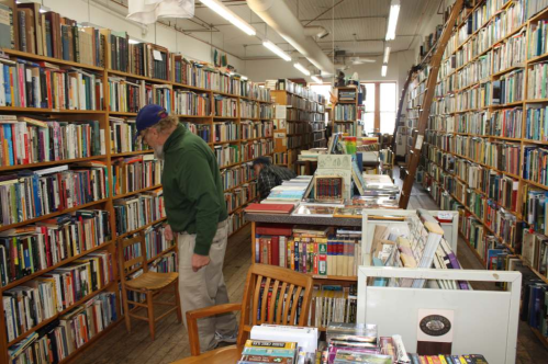 A cozy bookstore with tall shelves filled with books, featuring two people browsing and wooden chairs scattered around.