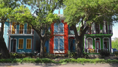 Colorful row of three houses with vibrant facades, surrounded by lush greenery and large trees.