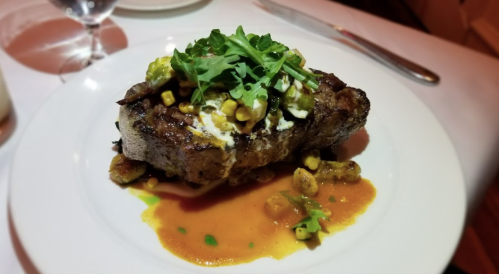 A beautifully plated steak topped with greens and corn, served with a flavorful sauce on a white dish.