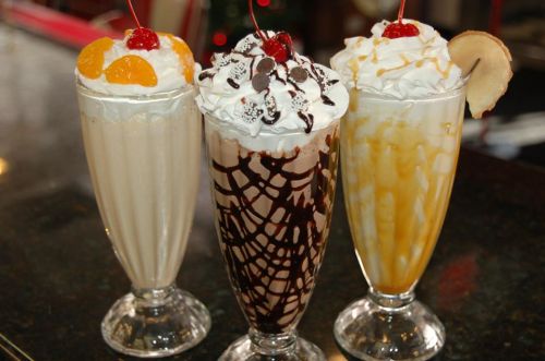 Three tall milkshakes topped with whipped cream, cherries, and various toppings: vanilla, chocolate, and caramel flavors.