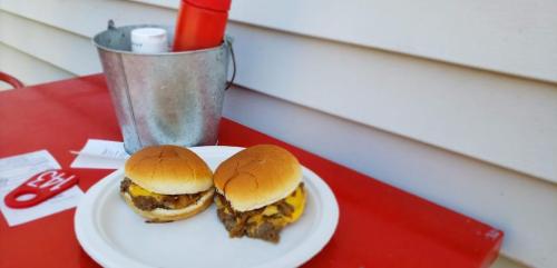 Two cheeseburgers on a white plate, with a red bucket and condiments in the background on a red table.