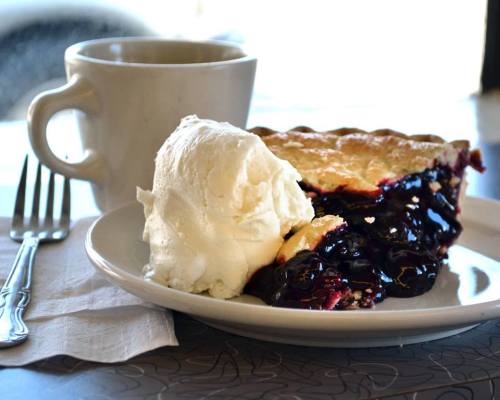 A slice of blueberry pie with a scoop of vanilla ice cream, served on a plate next to a cup of coffee.