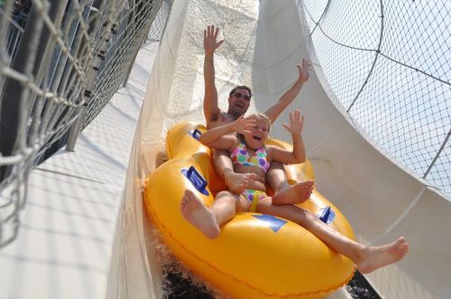 A man and a girl joyfully slide down a water slide on an inflatable tube, both with hands raised in excitement.