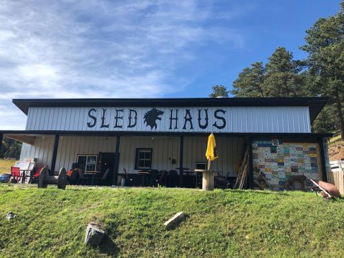 A rustic building with the sign "Sled Haus," surrounded by trees and outdoor seating.