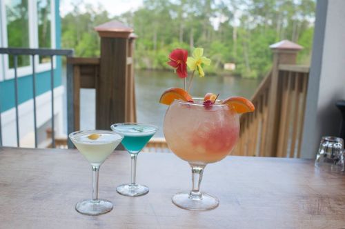 Three colorful cocktails on a wooden table, with a scenic view of trees and water in the background.