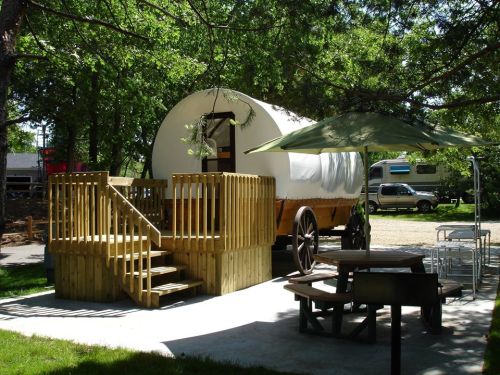 A wooden wagon with a white cover, surrounded by trees, featuring a deck, picnic table, and green umbrella.
