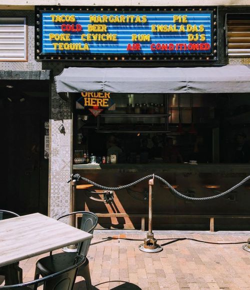 A colorful restaurant sign lists menu items like tacos, margaritas, and ceviche, with a shaded outdoor seating area.