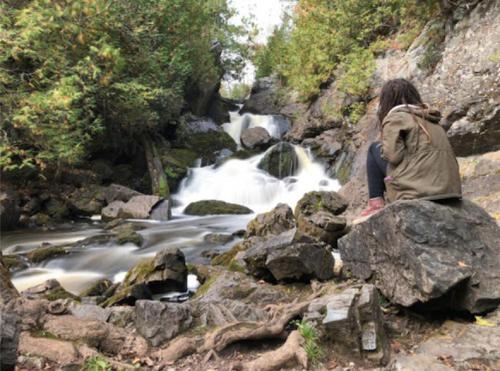 A person sits on a rock by a flowing waterfall, surrounded by lush greenery and rocky terrain.