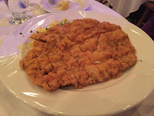 A large, golden-brown breaded and fried dish served on a white plate, garnished with lemon and parsley.