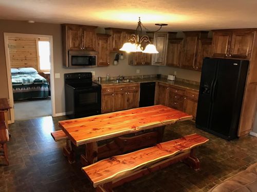 A rustic kitchen with wooden cabinets, a large dining table, and a view of a cozy bedroom in the background.