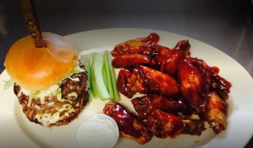 A plate featuring a stacked burger with lettuce and a side of saucy chicken wings and celery sticks.