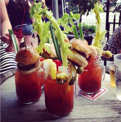 Three colorful cocktails in mason jars, garnished with celery, shrimp, and mini burgers, set on a wooden table.