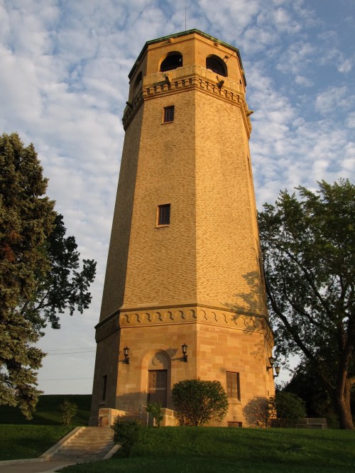 A tall, historic stone tower with a green roof, surrounded by trees and a blue sky with scattered clouds.