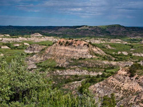 A panoramic view of rolling hills and layered rock formations under a cloudy sky, surrounded by greenery.