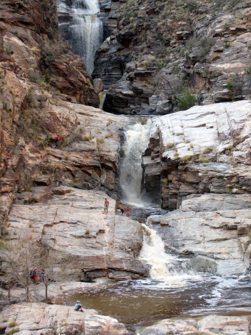 A scenic waterfall cascades down rocky cliffs, with hikers exploring the area around the water.