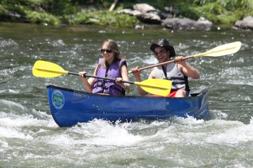 Two people paddle a blue canoe on a river, smiling and enjoying a sunny day.