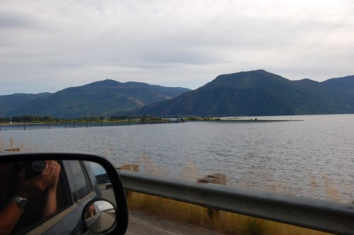 A scenic view of mountains and a calm lake, captured from a car window with a glimpse of the side mirror.