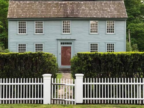 A blue two-story house with a shingled roof, surrounded by a green hedge and a white picket fence.