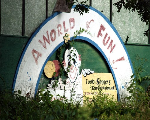A colorful, weathered sign featuring a clown and the text "A World of Fun!" surrounded by overgrown foliage.