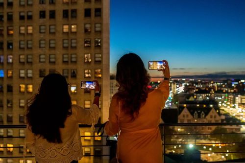 Two women stand on a balcony at dusk, taking photos of a city skyline illuminated by lights.