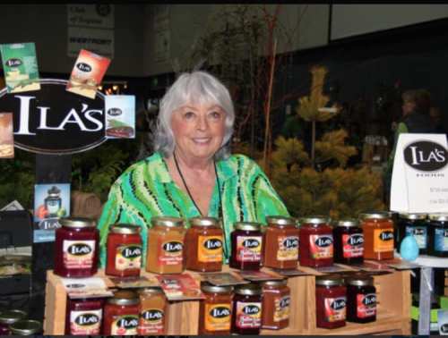 A smiling woman with gray hair stands behind a display of jars labeled "Ila's" at a market or fair.
