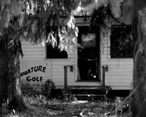 A black-and-white photo of a weathered building with a "Miniature Golf" sign, surrounded by trees.