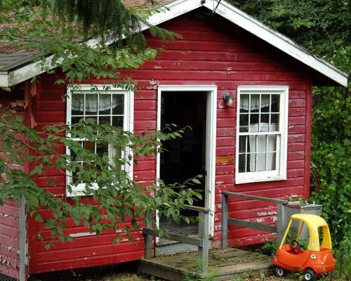 A small red wooden house surrounded by greenery, featuring a front porch and a yellow toy car nearby.