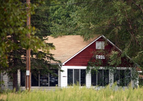 A red and white house partially obscured by trees, with a grassy foreground and power lines in the background.