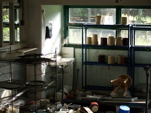 A cluttered room with shelves of pots, scattered debris, and sunlight streaming through the windows.