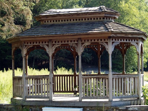 A wooden gazebo with a sloped roof, surrounded by greenery and set against a natural backdrop.