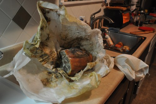 A burnt, charred object wrapped in stained paper sits on a kitchen counter near a sink and dirty dishes.