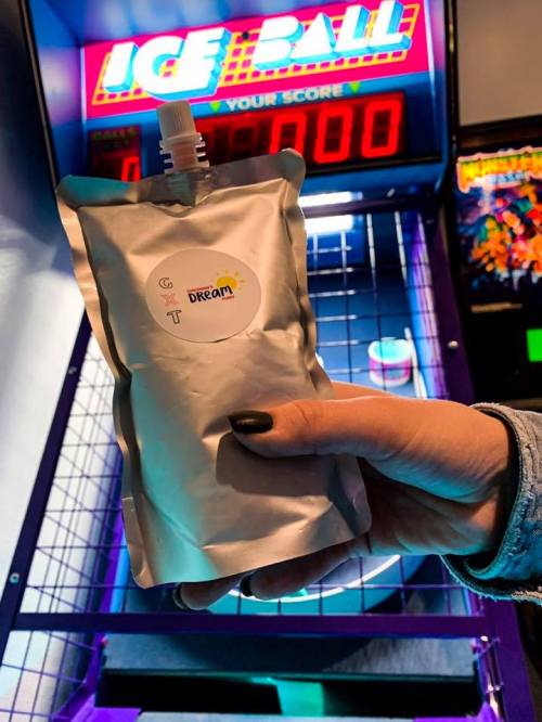 A hand holds a silver pouch labeled "Dream" in front of an arcade game with a score display.