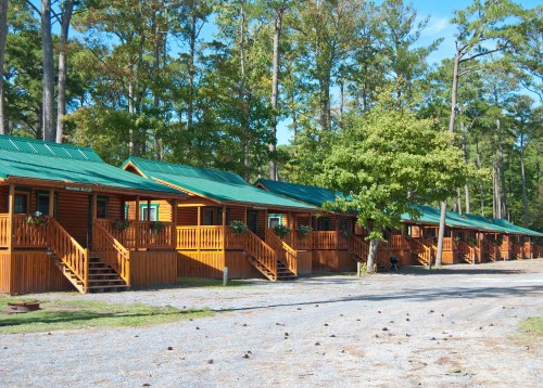 A row of wooden cabins with green roofs, surrounded by trees and a gravel path.