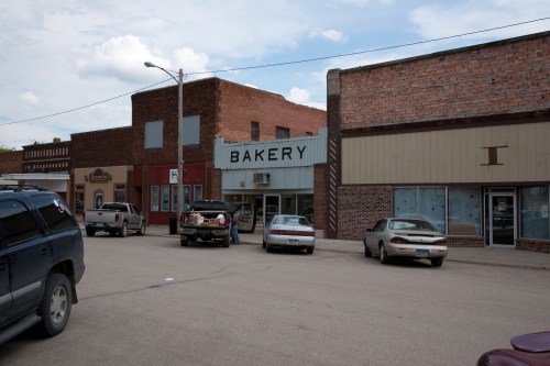 A small town street featuring a bakery sign, parked cars, and brick buildings under a cloudy sky.