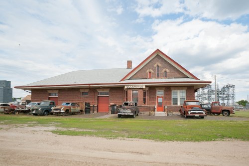 Historic brick train depot with vintage cars parked outside, set against a cloudy sky. Green grass in the foreground.