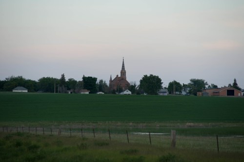 A distant view of a church steeple surrounded by trees and houses, set against a green field under a soft sky.