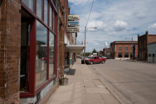 A quiet street scene featuring an insurance agency, parked cars, and a water tower in the background under a cloudy sky.