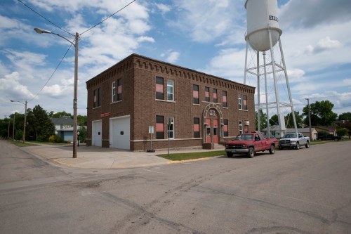 A brick building with red accents and a water tower nearby, set on a quiet street under a partly cloudy sky.