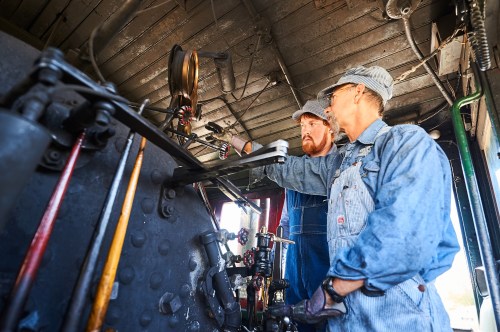 Two men in overalls work together inside a vintage steam locomotive, adjusting controls and machinery.