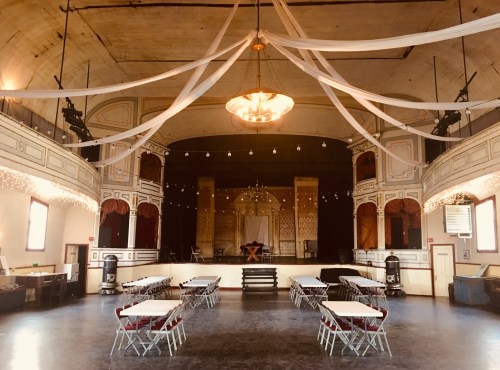 A spacious event hall with tables set up, draped fabric overhead, and stage lights creating a festive atmosphere.