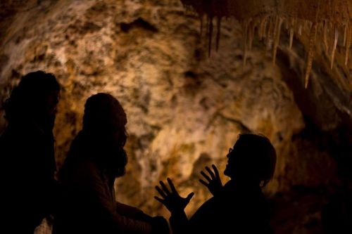 Silhouettes of three people in a cave, with stalactites hanging above and warm lighting illuminating the rocky background.
