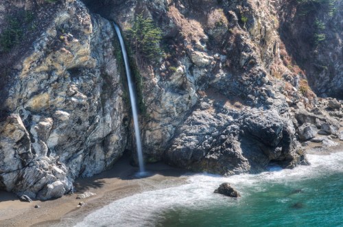 A serene coastal scene featuring a waterfall cascading down rocky cliffs into a sandy beach and turquoise ocean.