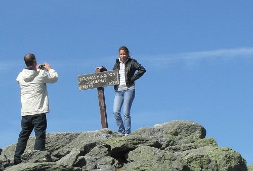 A woman stands on a rock at the summit of Mount Washington, while a man takes her photo beside the summit sign.