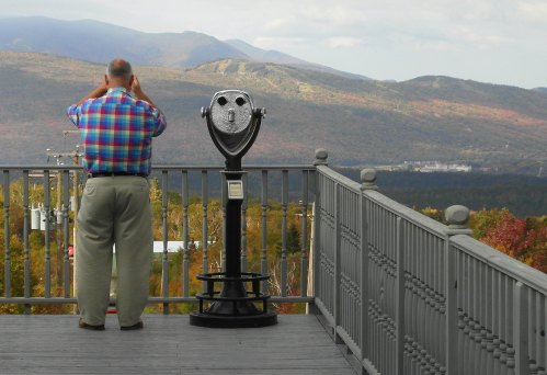 A man in a plaid shirt looks through a binocular viewer at a scenic mountain landscape.